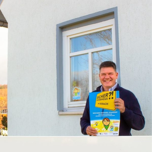 Bürgermeister Gerrit Häcker steht mit einem Notinsel Plakat in der Hand vor dem Gebäude der Gemeinde Seebach.